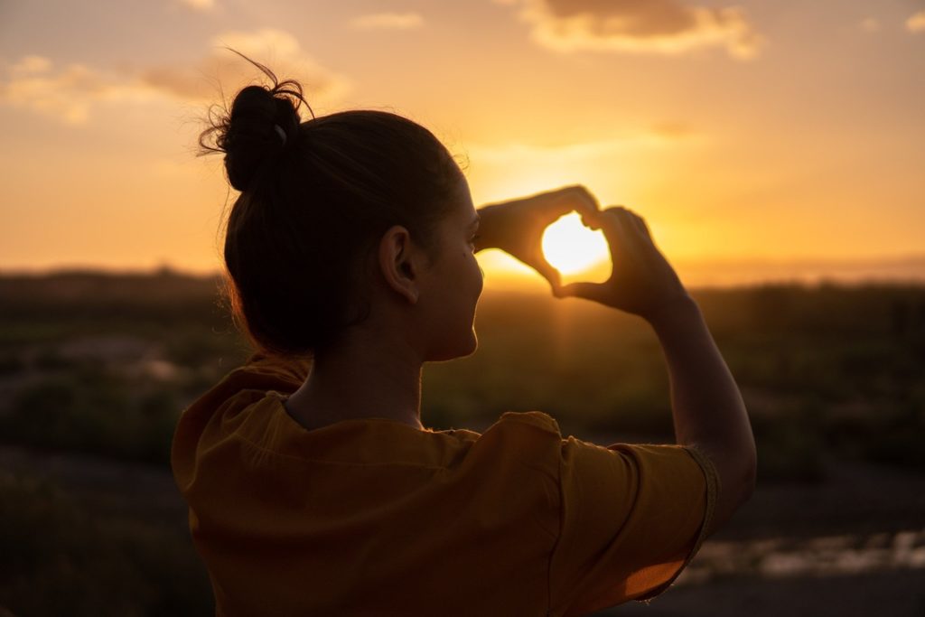 una ragazza fa il simbolo del cuore con le mani guardando il tramonto