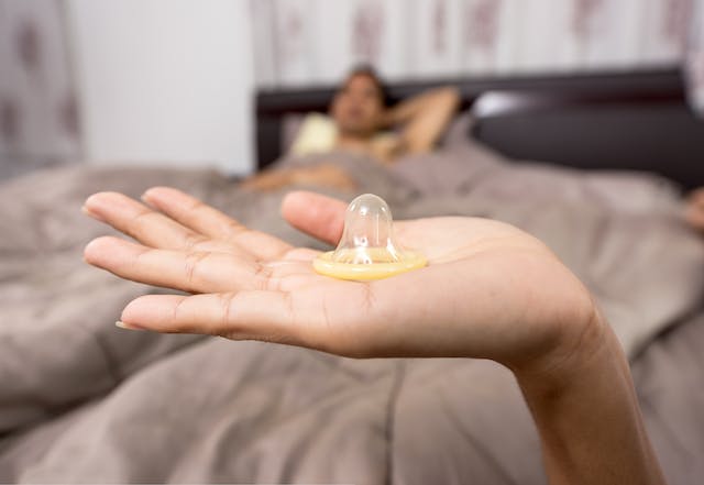 una mano mostra un preservativo aperto con un uomo che aspetta a letto sullo sfondo